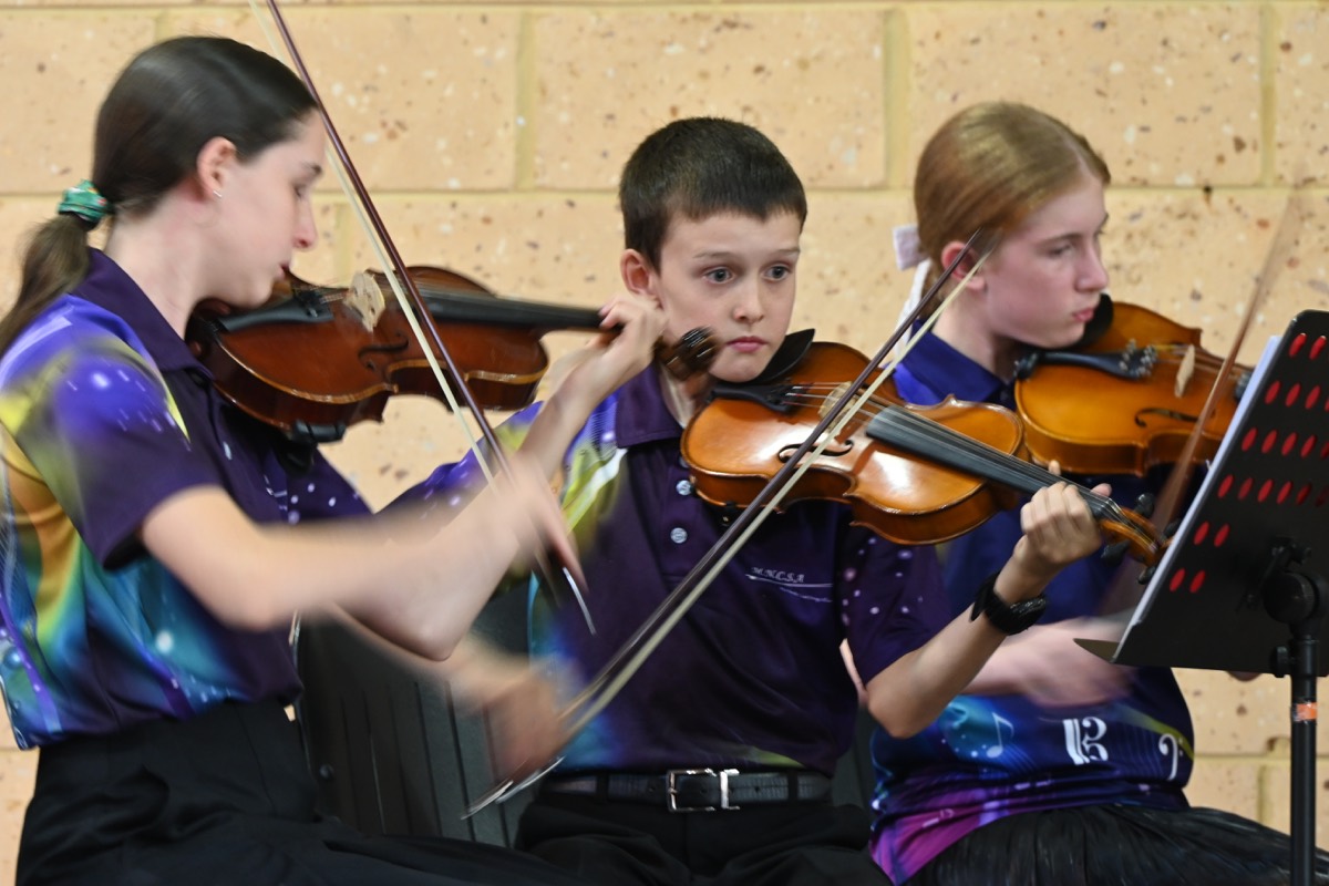 Three young violinists in MNCYO shirts performing together at the 2025 concert.