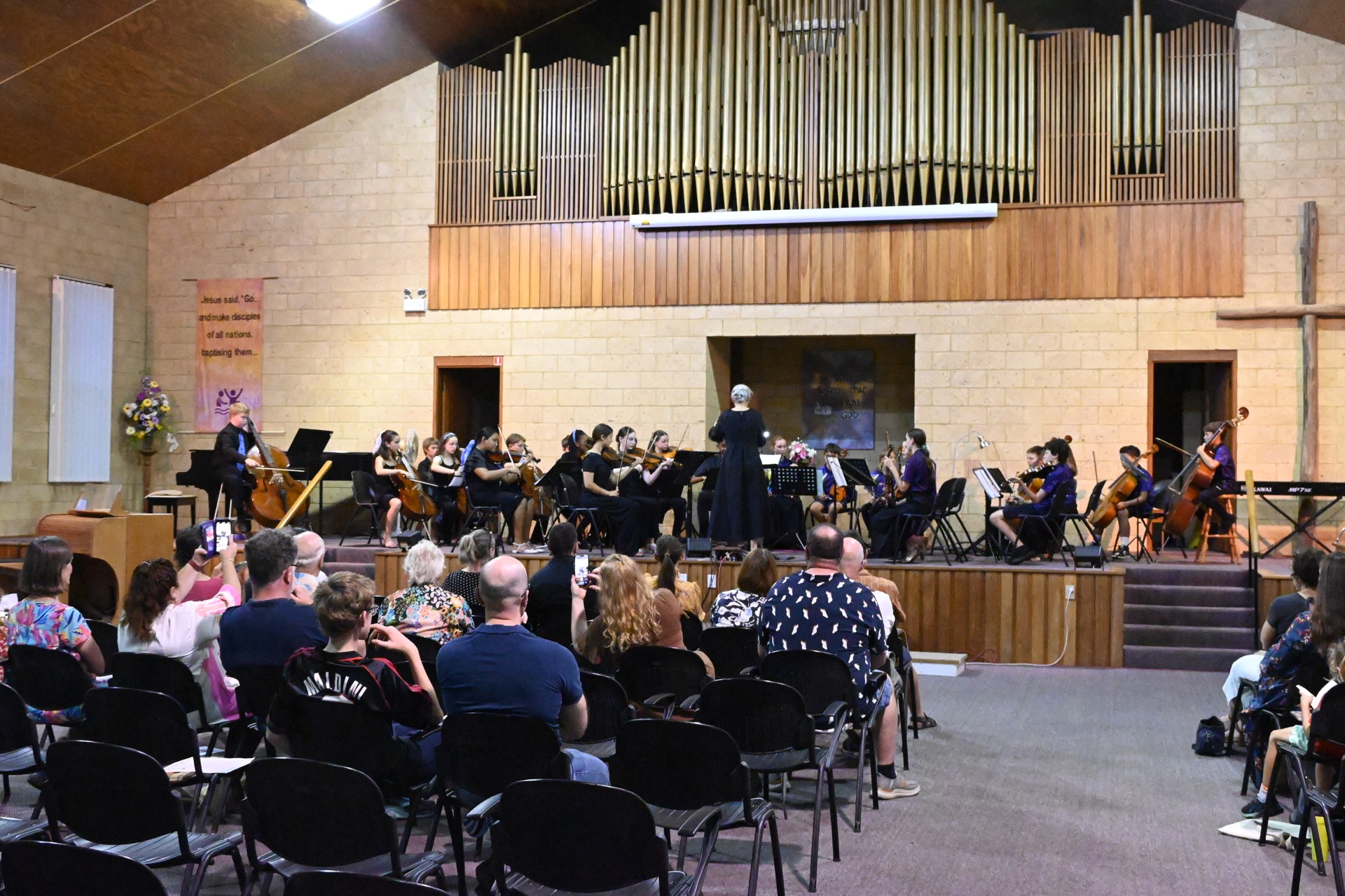 MNCYO performing on stage with conductor, strings, piano, and audience watching.